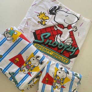 May include: A white t-shirt featuring a graphic of Snoopy and Woodstock with the text "Snoopy SPIRIT SPIRIT" and two folded white towels with blue stripes, yellow triangles, and graphics of Snoopy and Woodstock playing sports.