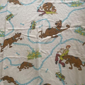 May include: White fabric with a repeating pattern of Scooby Doo and Shaggy running and driving the Mystery Machine. The background is a light blue with a chain link pattern.