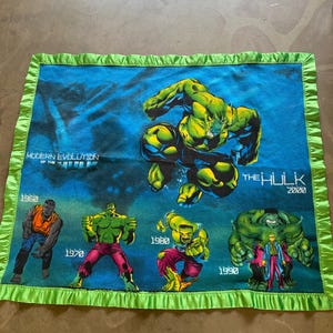 May include: A rectangular blue fleece blanket with a green satin ruffled border featuring graphic illustrations of The Hulk from different decades. The text reads Modern Evolution of the Hulk, 1960, 1970, 1980, 1990, and The Hulk 2000.