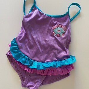May include: Purple swimsuit with a blue and pink ruffle trim. The swimsuit has a Cabbage Patch Kids logo on the front.