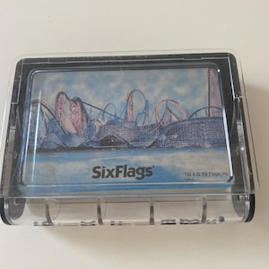 May include: A clear plastic souvenir featuring a roller coaster scene with blue, red, and gray elements. The Six Flags logo is visible at the bottom of the image.