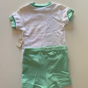 May include: A white short-sleeved t-shirt with green trim around the collar and sleeves. The t-shirt is paired with green shorts. The tag says "Size 18M".
