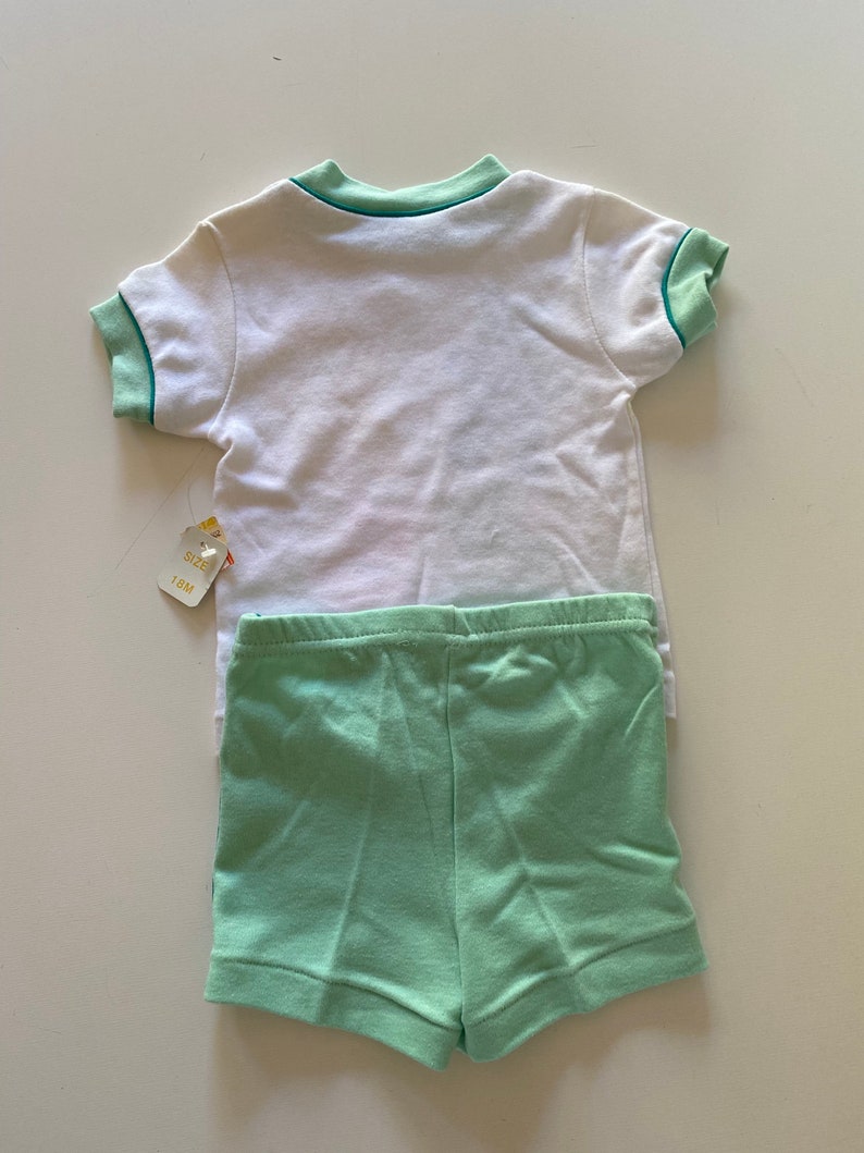 May include: A white short-sleeved t-shirt with green trim around the collar and sleeves. The t-shirt is paired with green shorts. The tag says "Size 18M".