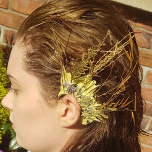 May include: A woman with long brown hair wearing a gray shirt with a floral ear cuff made of dried flowers and grasses.