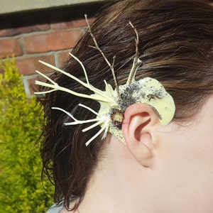May include: A close-up of a person's ear with a handmade ear cuff made of twigs and a light yellow material. The cuff is attached to the ear with a small piece of tape.