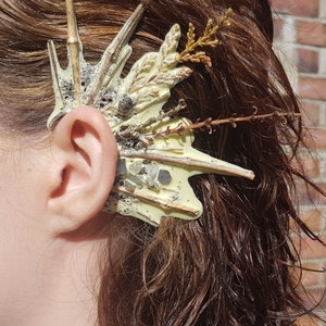 May include: A white ear cuff with a textured surface and natural elements, including twigs and dried flowers, attached to it.