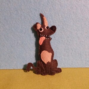 Könnte beinhalten: Handgefertigte Filzhundefigur in sitzender Position. Der Hund ist hauptsächlich braun mit einem hellrosa Bauch und Zunge. Er hat große, runde Augen und einen kleinen, gebogenen Schwanz. Der Hintergrund ist hellblau und gelb.