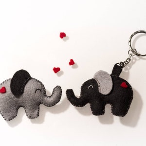 May include: Two felt elephant keychains, one gray and one black, with red felt hearts on them. The black elephant is attached to a silver key ring. The elephants are facing each other and there are small red felt hearts scattered around them.