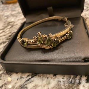 Puede incluir: Una pulsera dorada con bisagras y dise&ntilde;o de corona. La corona est&aacute; adornada con piedras preciosas verdes ovaladas y peque&ntilde;as perlas. La pulsera se exhibe en una caja de joyer&iacute;a negra con un fondo de m&aacute;rmol gris.