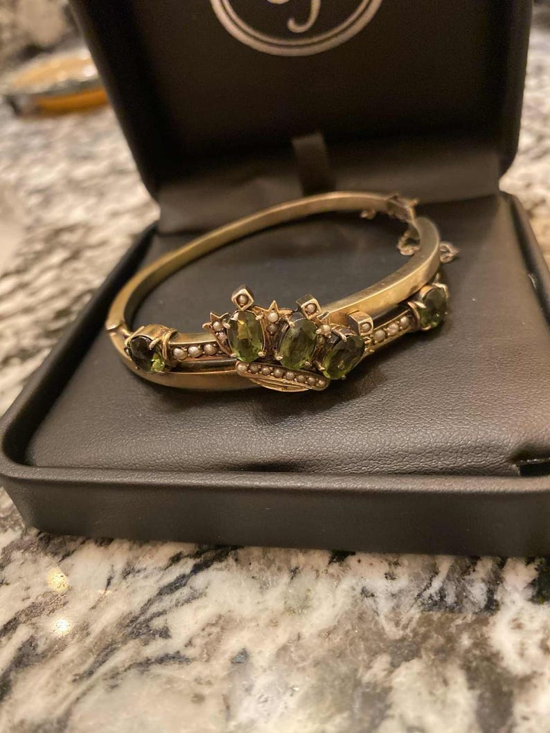Puede incluir: Una pulsera dorada con bisagras y dise&ntilde;o de corona. La corona est&aacute; adornada con piedras preciosas verdes ovaladas y peque&ntilde;as perlas. La pulsera se exhibe en una caja de joyer&iacute;a negra con un fondo de m&aacute;rmol gris.