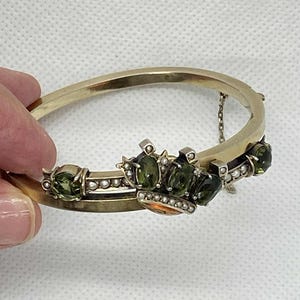 Puede incluir: Una pulsera dorada con bisagras y dise&ntilde;o de corona. La corona presenta piedras preciosas verdes y peque&ntilde;as perlas. La pulsera est&aacute; sujeta por una mano sobre un fondo blanco. La pulsera tiene una peque&ntilde;a cadena para mayor seguridad.