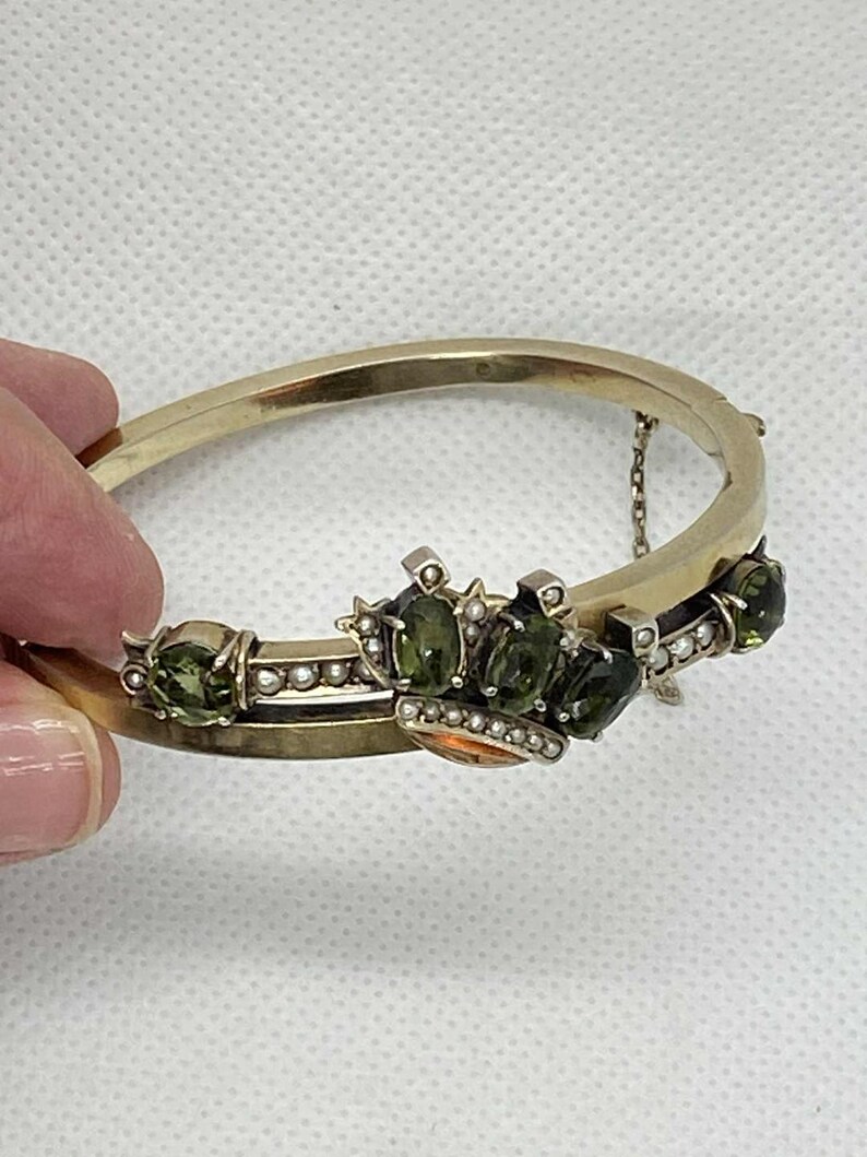 Puede incluir: Una pulsera dorada con bisagras y dise&ntilde;o de corona. La corona presenta piedras preciosas verdes y peque&ntilde;as perlas. La pulsera est&aacute; sujeta por una mano sobre un fondo blanco. La pulsera tiene una peque&ntilde;a cadena para mayor seguridad.