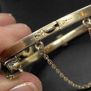 Puede incluir: Primer plano de una pulsera de brazalete dorada con bisagras. La pulsera presenta una cadena de seguridad y un cierre decorativo con detalles grabados. Se ve una peque&ntilde;a piedra preciosa de talla cuadrada.
