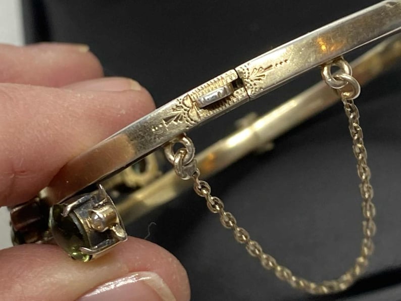 Puede incluir: Primer plano de una pulsera de brazalete dorada con bisagras. La pulsera presenta una cadena de seguridad y un cierre decorativo con detalles grabados. Se ve una peque&ntilde;a piedra preciosa de talla cuadrada.