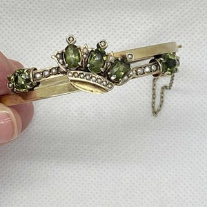 Puede incluir: Una pulsera dorada con un dise&ntilde;o de corona, con piedras preciosas de color verde oliva y peque&ntilde;os detalles de perlas. La pulsera tiene un cierre de bisagra y una delicada cadena. Las piedras preciosas son de forma ovalada y est&aacute;n dispuestas en un patr&oacute;n decorativo.