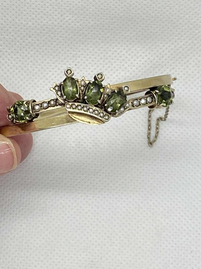 Puede incluir: Una pulsera dorada con un dise&ntilde;o de corona, con piedras preciosas de color verde oliva y peque&ntilde;os detalles de perlas. La pulsera tiene un cierre de bisagra y una delicada cadena. Las piedras preciosas son de forma ovalada y est&aacute;n dispuestas en un patr&oacute;n decorativo.