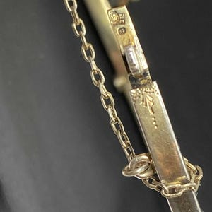 Puede incluir: Primer plano de una pulsera dorada con una delicada cadena y cierre. La pulsera presenta detalles y marcas intrincadas, lo que sugiere que es vintage o antigua. El fondo oscuro resalta la joya.