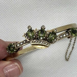 Puede incluir: Pulsera dorada con dise&ntilde;o de corona y tres piedras preciosas ovaladas de color verde oliva. La corona est&aacute; adornada con peque&ntilde;as perlas. La pulsera tiene un dise&ntilde;o de bisagra y una delicada cadena.