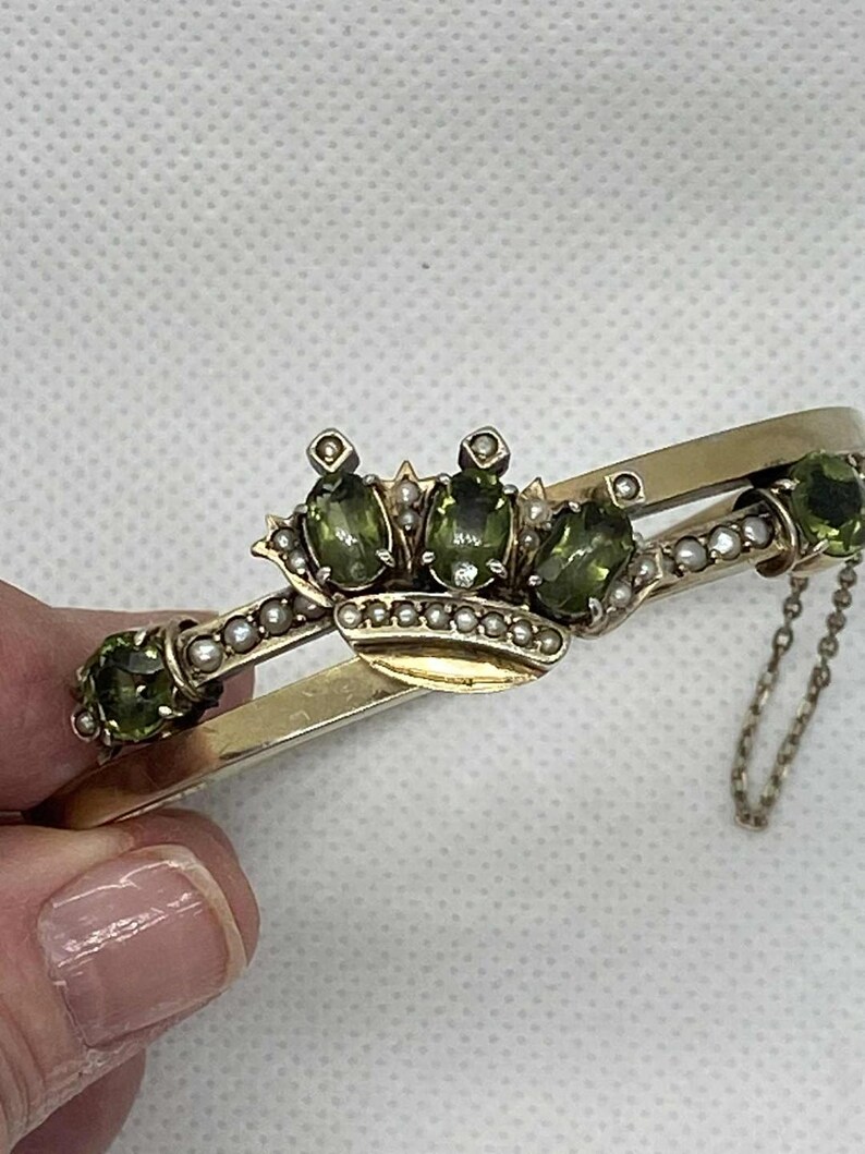 Puede incluir: Pulsera dorada con dise&ntilde;o de corona y tres piedras preciosas ovaladas de color verde oliva. La corona est&aacute; adornada con peque&ntilde;as perlas. La pulsera tiene un dise&ntilde;o de bisagra y una delicada cadena.