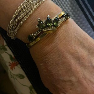 Puede incluir: Una pulsera de plata con pedrer&iacute;a y una pulsera dorada con un dise&ntilde;o de corona. La corona presenta piedras preciosas verdes y peque&ntilde;as perlas blancas. Las pulseras se llevan en una mu&ntilde;eca.