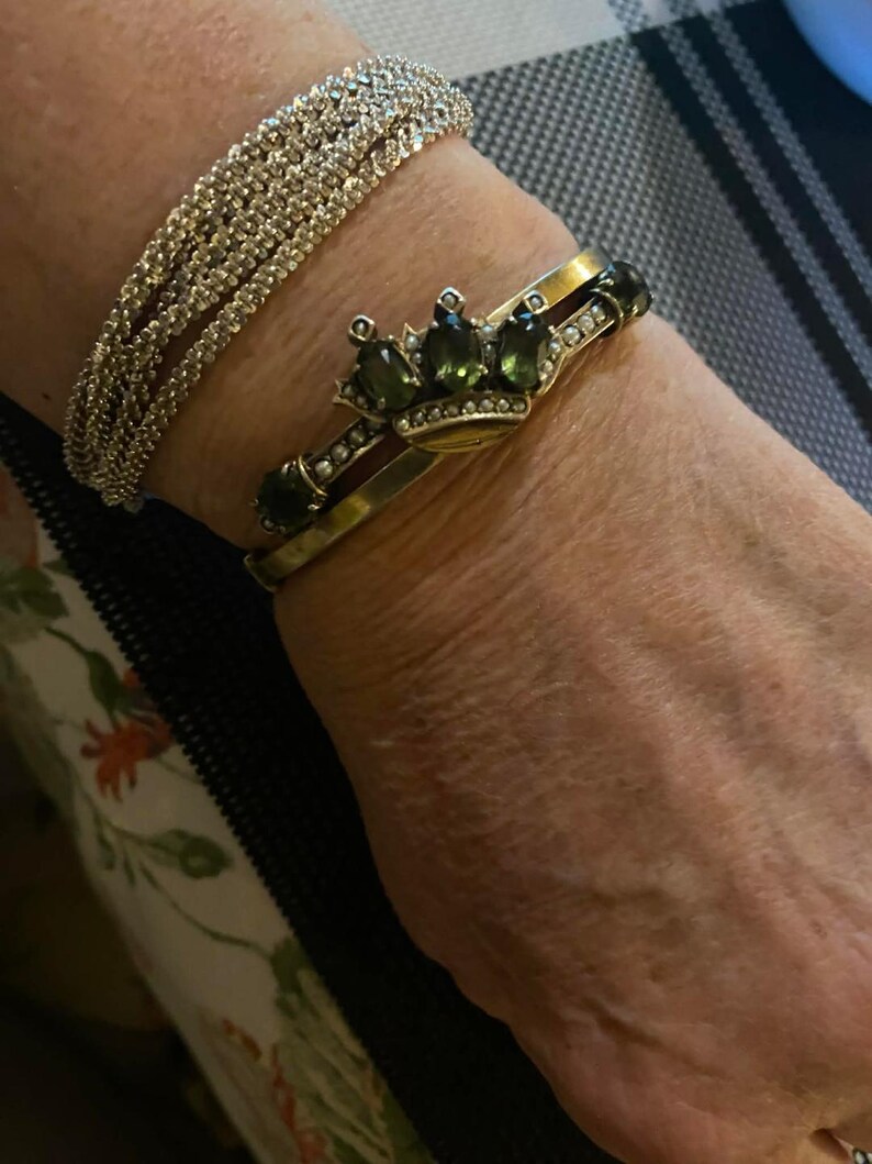 Puede incluir: Una pulsera de plata con pedrer&iacute;a y una pulsera dorada con un dise&ntilde;o de corona. La corona presenta piedras preciosas verdes y peque&ntilde;as perlas blancas. Las pulseras se llevan en una mu&ntilde;eca.