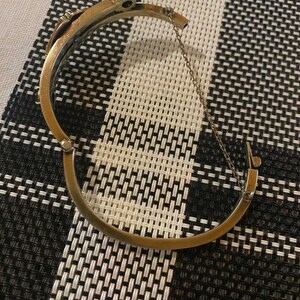 Puede incluir: Una pulsera de brazalete dorada con bisagras y una secci&oacute;n superior decorativa. La pulsera est&aacute; abierta, revelando el cierre y una cadena de seguridad. El fondo es un patr&oacute;n de cuadros blanco y negro.
