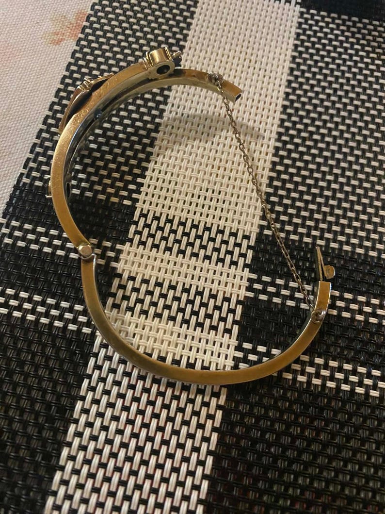 Puede incluir: Una pulsera de brazalete dorada con bisagras y una secci&oacute;n superior decorativa. La pulsera est&aacute; abierta, revelando el cierre y una cadena de seguridad. El fondo es un patr&oacute;n de cuadros blanco y negro.
