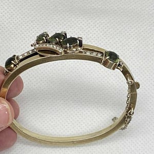 Puede incluir: Pulsera dorada con bisagras con piedras preciosas verdes ovaladas y peque&ntilde;as perlas. La pulsera presenta una pieza central decorativa con m&uacute;ltiples piedras preciosas y un delicado cierre de cadena. Una joya cl&aacute;sica.