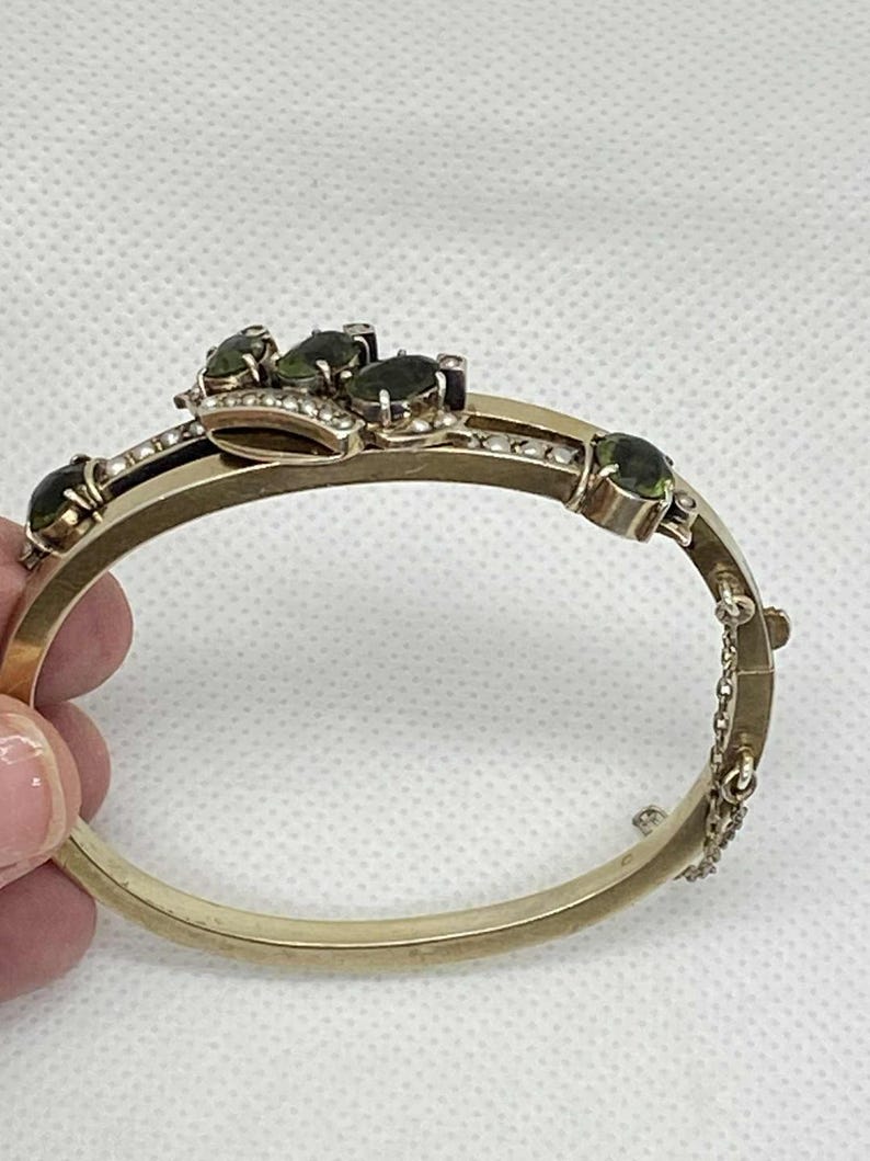 Puede incluir: Pulsera dorada con bisagras con piedras preciosas verdes ovaladas y peque&ntilde;as perlas. La pulsera presenta una pieza central decorativa con m&uacute;ltiples piedras preciosas y un delicado cierre de cadena. Una joya cl&aacute;sica.