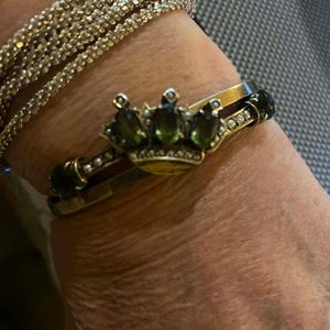 Puede incluir: Un brazalete dorado con un dise&ntilde;o de corona con piedras preciosas verdes y un brazalete de pedrer&iacute;a. El brazalete de corona tiene una banda dorada con piedras verdes y peque&ntilde;as perlas. El brazalete de pedrer&iacute;a est&aacute; hecho de m&uacute;ltiples hilos de peque&ntilde;os brillantes transparentes.