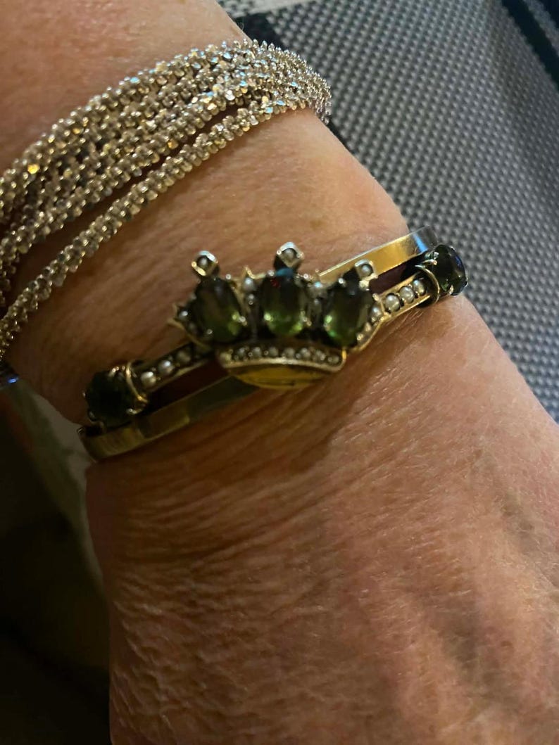 Puede incluir: Un brazalete dorado con un dise&ntilde;o de corona con piedras preciosas verdes y un brazalete de pedrer&iacute;a. El brazalete de corona tiene una banda dorada con piedras verdes y peque&ntilde;as perlas. El brazalete de pedrer&iacute;a est&aacute; hecho de m&uacute;ltiples hilos de peque&ntilde;os brillantes transparentes.