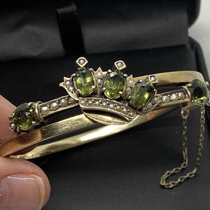 Puede incluir: Un brazalete dorado con un dise&ntilde;o de bisagra, con piedras preciosas verdes ovaladas y peque&ntilde;as perlas. El brazalete tiene un elemento decorativo en forma de corona y una delicada cadena. El fondo es una caja de joyer&iacute;a negra.