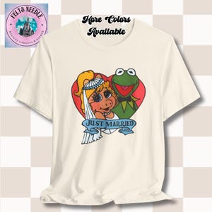 May include: Cream-colored t-shirt featuring a heart-shaped graphic with Miss Piggy in a bridal veil and Kermit the Frog. The graphic includes the text "Just Married" in a blue banner. The shirt also has the text "More Colors Available".