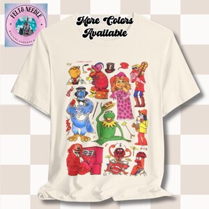 May include: White t-shirt featuring a colourful graphic print of characters from the Muppets. The characters include Kermit the Frog, Miss Piggy, Animal, and Gonzo. The text "More Colors Available" is printed at the top of the shirt.