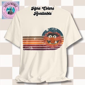 Könnte beinhalten: Ein weißes T-Shirt mit einem Vintage-Grafikdruck eines roten und orangefarbenen Muppet-Charakters vor blauem Hintergrund. Die Grafik hat einen abgenutzten Look mit verblassten Farben und einem gestreiften Design. Der Text "Mehr Farben Verfügbar" ist in einer Retro-Schriftart oben auf dem Shirt gedruckt.