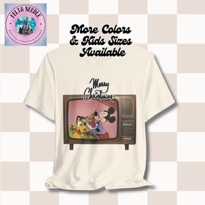 May include: A white t-shirt featuring a vintage television screen graphic with Mickey Mouse and Pluto. The television screen shows Mickey Mouse and Pluto decorating a Christmas tree. The text "Merry Christmas" is above the television screen. The text "More Colors & Kids Sizes Available" is at the top of the image.