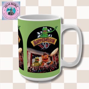 May include: White ceramic mug featuring a green background with a Muppet Vision 3D design. The design includes Kermit the Frog, Fozzie Bear, and the Muppet Vision 3D logo. The mug has a large handle and is suitable for hot or cold beverages.