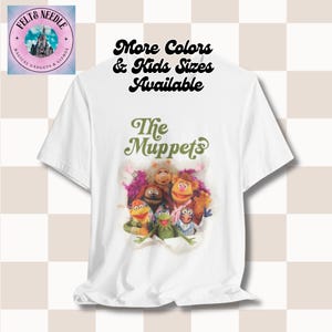 May include: White t-shirt with a green and white graphic of the Muppets characters. The text "The Muppets" is above the image. The text "More Colors & Kids Sizes Available" is above the image.