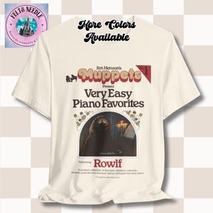 May include: Cream-colored t-shirt featuring a graphic from Jim Henson's Muppets. The design includes the text "Muppets," "Very Easy Piano Favorites," and "Rowlf." The shirt also has the text "More Colors Available" at the top.