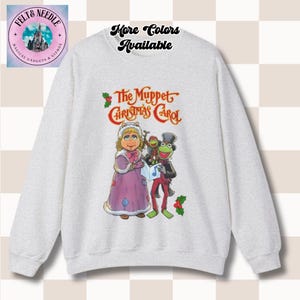 May include: A light gray sweatshirt featuring a graphic from "The Muppet Christmas Carol." The design includes Miss Piggy, Kermit the Frog, and other characters. The text "The Muppet Christmas Carol" is displayed in red and gold.