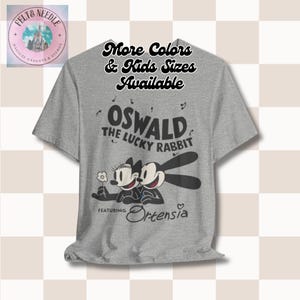 Può includere: Una maglietta grigia con Oswald il coniglio fortunato e Ortensia. Il design include il testo "Oswald The Lucky Rabbit" e "Featuring Ortensia". La maglietta ha anche il testo "More Colors & Kids Sizes Available".