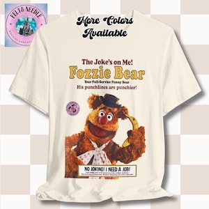 May include: Cream-colored t-shirt featuring a graphic of Fozzie Bear holding a banana, with text that reads "The Joke's on Me! Fozzie Bear." The shirt also includes the text "More Colors Available."