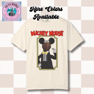 May include: A cream-colored short-sleeve t-shirt featuring a graphic of a puppet-style Mickey Mouse character in a black tuxedo with a yellow bow tie, framed in a gold border. The text MICKEY MOUSE appears in red above the frame.