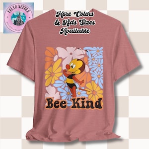 May include: A pink t-shirt with a cartoon bee and flowers printed on the front. The text "Bee Kind" is printed below the bee. The text "More Colors & Kids Sizes Available" is printed on the top of the shirt.
