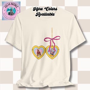 Könnte beinhalten: Ein weißes T-Shirt mit einem goldenen Herzmedaillon mit einer rosa Schleife. Das Medaillon enthält zwei Bilder, eines von einem roten Hahn und eines von einem blauen Monster. Der Text "Mehr Farben verfügbar" ist auf dem T-Shirt gedruckt.
