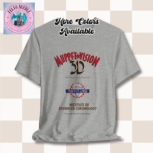 Może przedstawiać: Szara koszulka z napisem "Muppet Vision 3D" w kolorze czerwonym i złotym, wraz z innymi tekstami i logo. Koszulka ma również napis "More Colors Available" w kursywie. Koszulka jest od Felt & Needle.