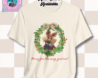 May include: Cream-colored t-shirt featuring a Muppet character inside a green wreath with red ribbon accents. The text "Penny for the song, guv'nor?" is printed below the wreath. The shirt also has the text "More Colors Available".