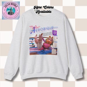 May include: A light gray sweatshirt featuring a graphic of Miss Piggy in an aerobics pose. The graphic includes the text "Miss Piggy's Aerobique" in blue and pink. The sweatshirt also has the text "More Colors Available" in black.