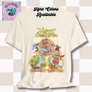 May include: Cream-coloured t-shirt featuring a graphic of Muppet characters from the Muppet Classic Theater. The design includes Miss Piggy, Kermit the Frog, and Fozzie Bear. Text on the shirt reads "More Colors Available" and "Muppet Classic Theater."