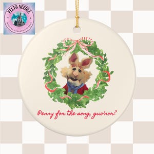 May include: A round, off-white ceramic ornament featuring a Muppet character inside a green wreath with red ribbon. The character is wearing a red jacket and blue scarf. The text "Penny for the song, guv'non?" is written in red.